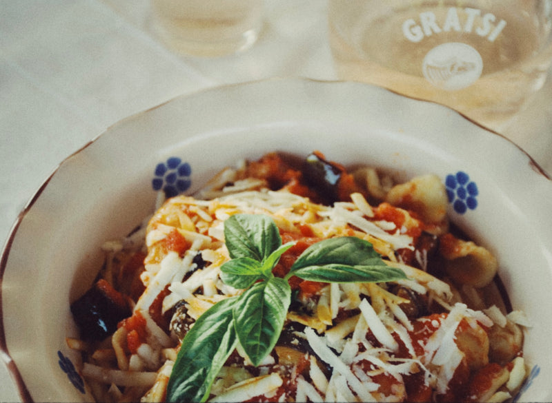A bowl of pasta topped with tomato sauce, grated cheese, and fresh basil leaves sits on a white table next to a glass with GRATSI printed on it.