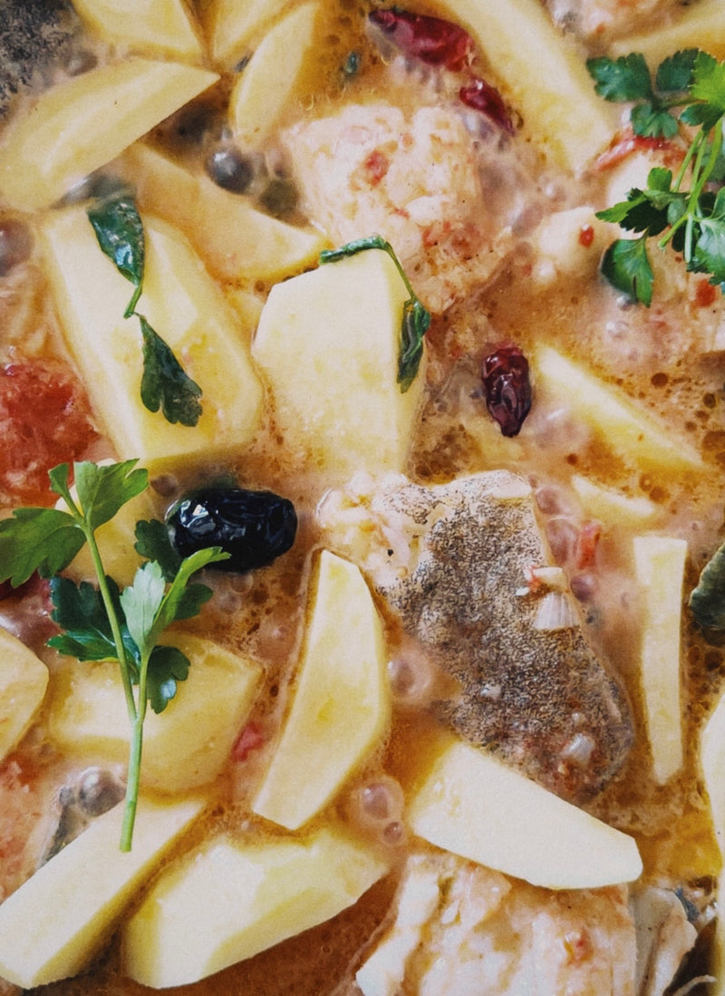 A close-up of a simmering stew with pieces of white fish, potato slices, parsley leaves, black olives, red chilies, and a light orange-colored broth.