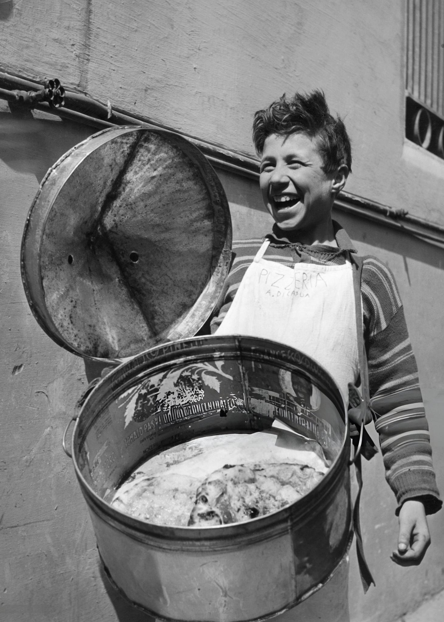 A smiling boy wearing an apron holds a large round metal container with a lid, displaying several pizzas inside, standing against a wall outdoors.