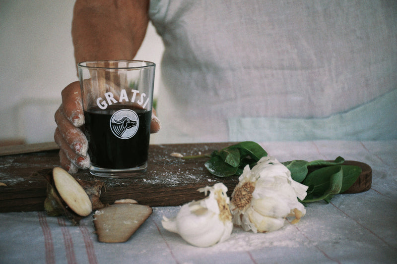A person with flour-dusted hands holds a glass of dark liquid labeled GRATS. On the table are garlic bulbs, leafy greens, a potato, and a wooden board, evoking a kitchen or cooking scene.