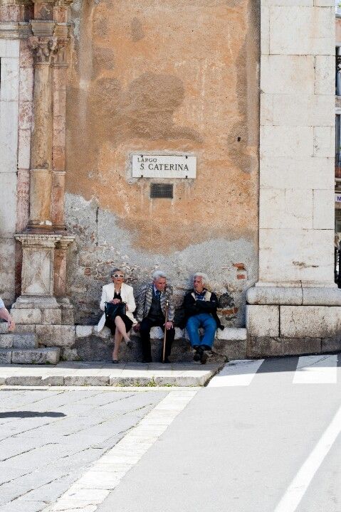 Three elderly people sit on a bench against a weathered wall beneath a street sign that reads Piazza S. Caterina on a sunny day. The wall is old with peeling paint and stone detailing.