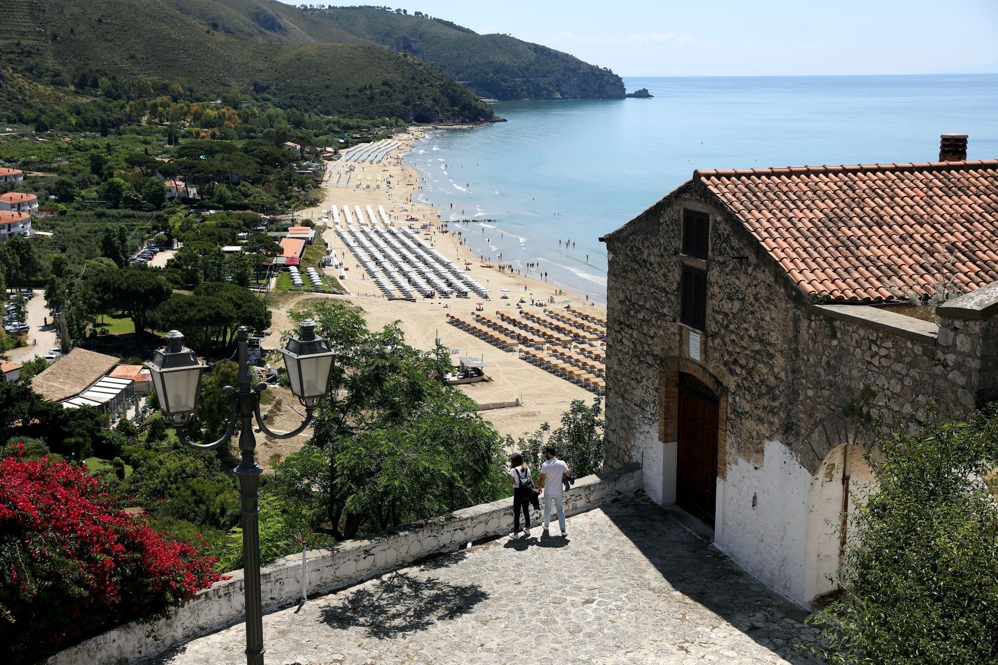A seaside village overlooks a sandy beach lined with umbrellas and sunbathers, surrounded by green hills and calm blue water. An old stone building with a red-tiled roof and a lamppost are in the foreground.
