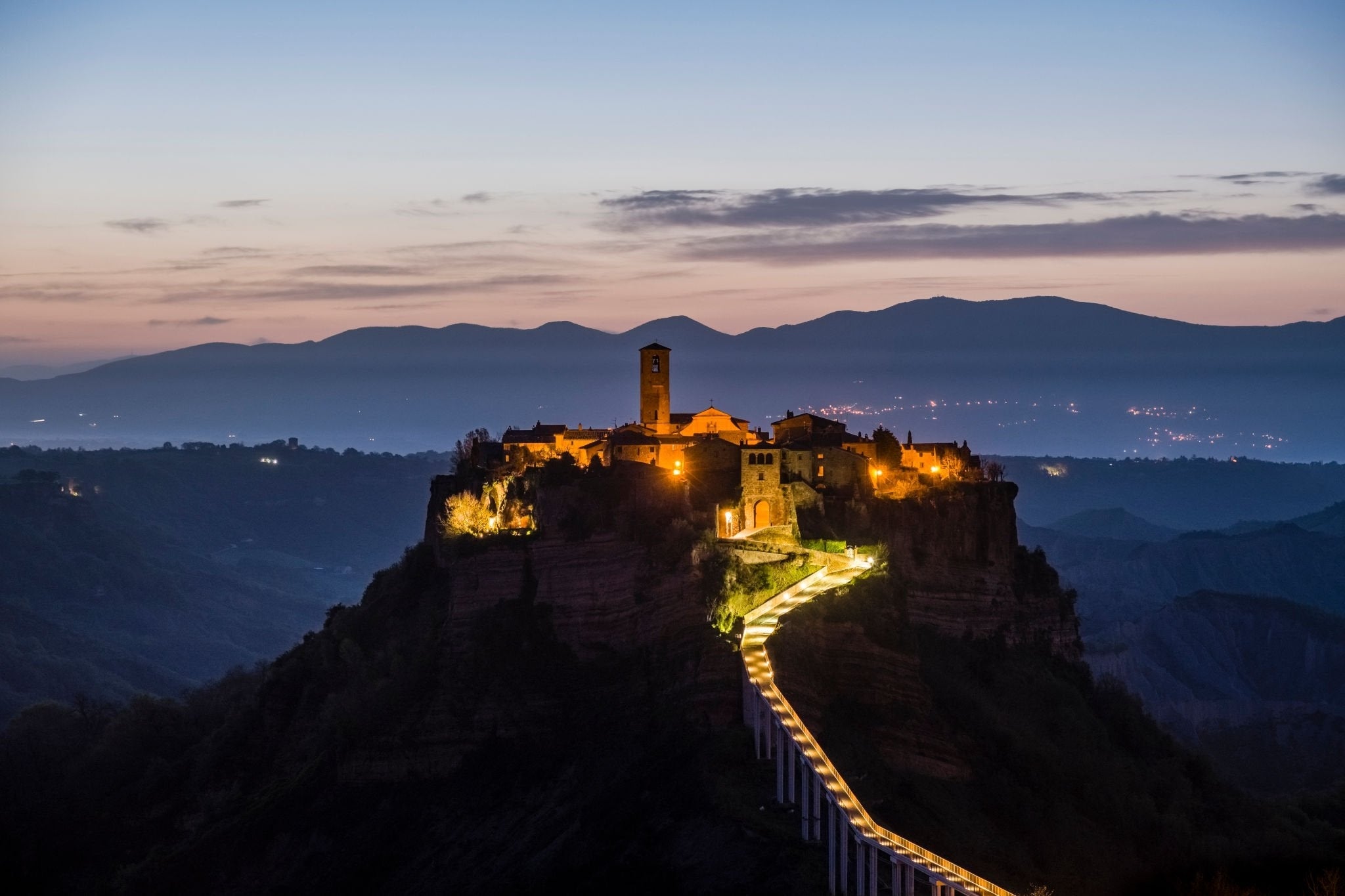A hilltop village, illuminated by warm lights, sits atop a cliff at dusk with mountains in the background and a winding, lit-up walkway leading to it.