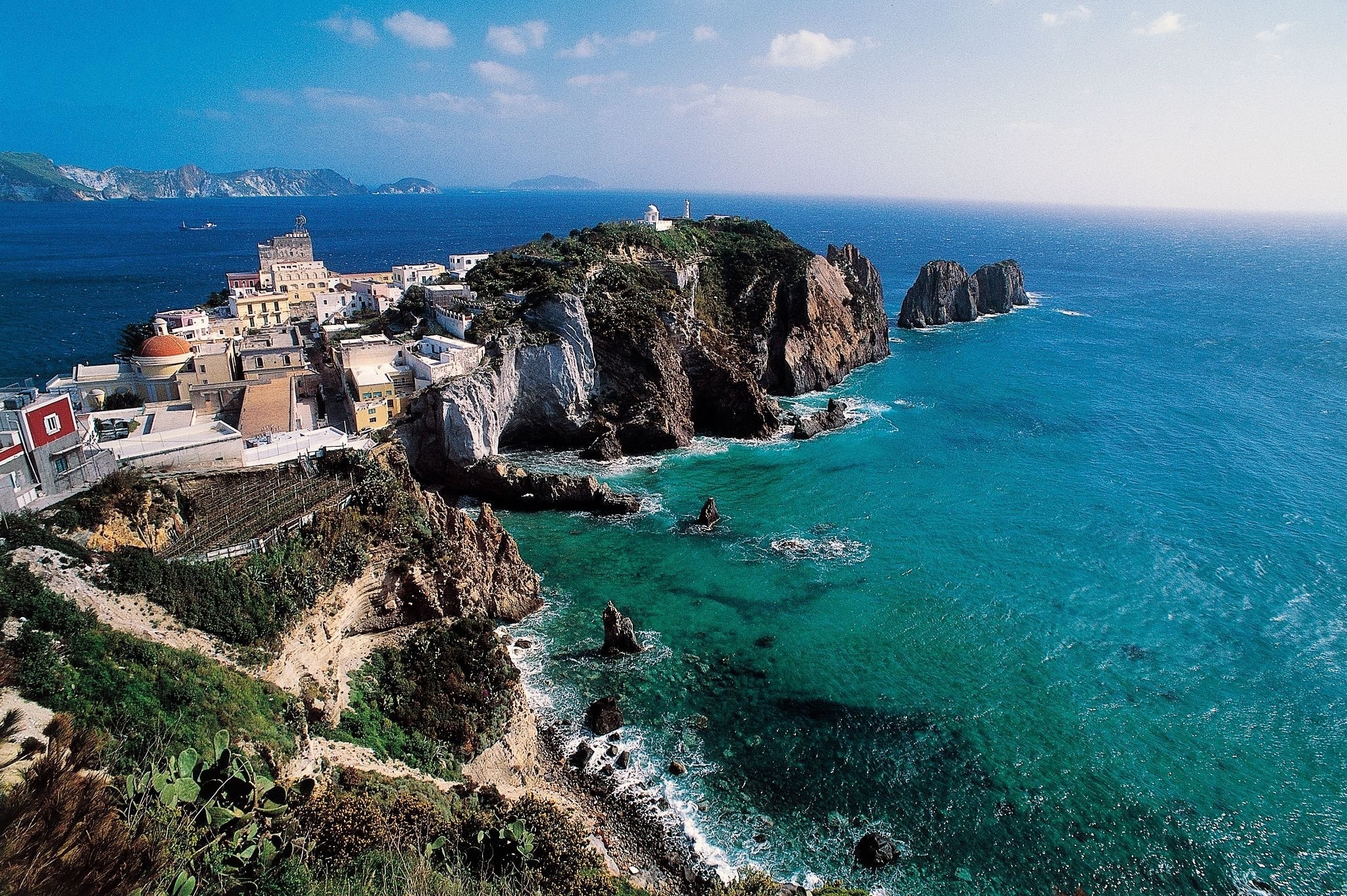 A coastal village with colorful buildings sits atop rocky cliffs overlooking clear turquoise sea, with waves gently crashing below and a lighthouse visible in the distance under a bright blue sky.