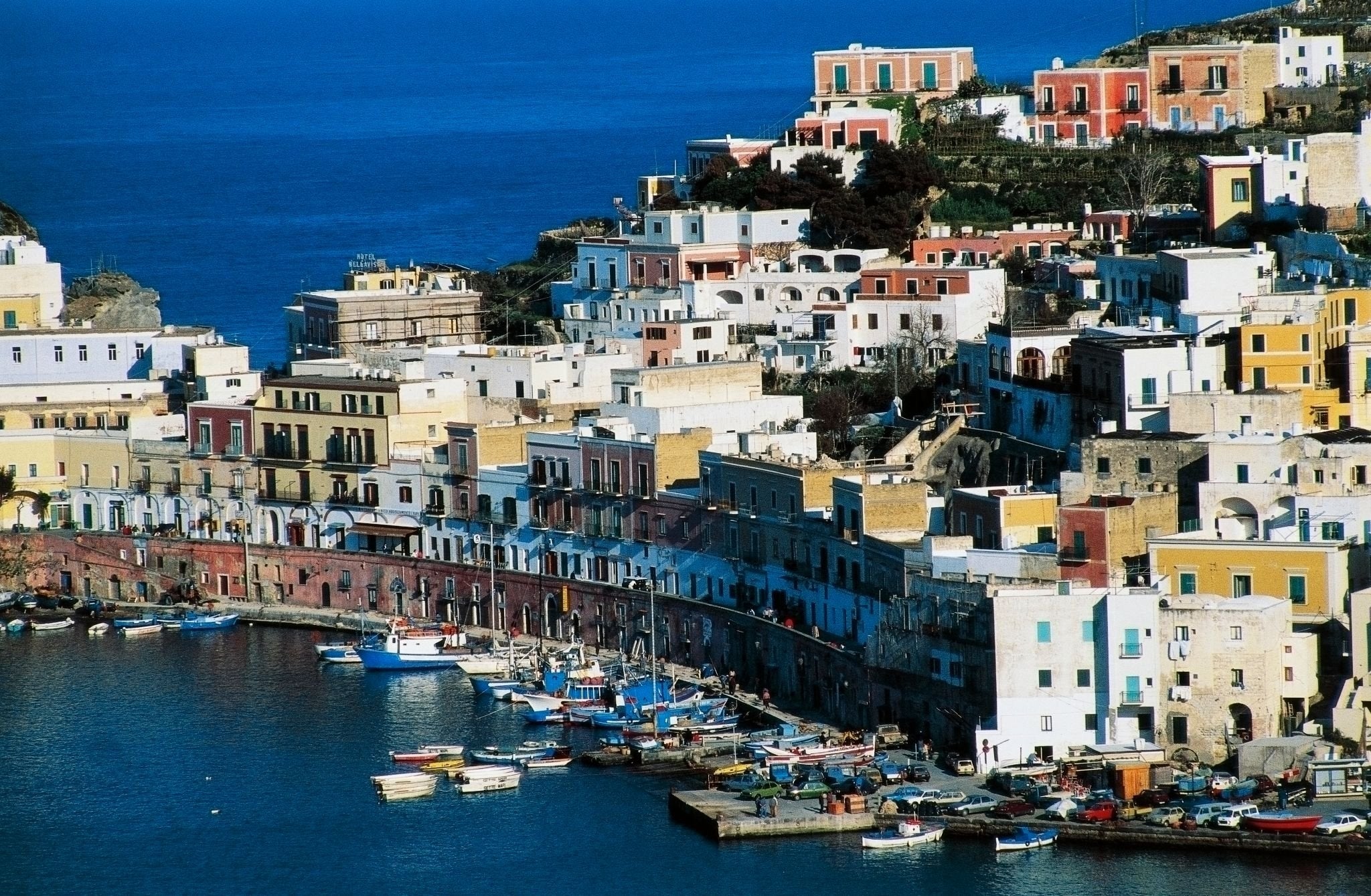 A seaside town with colorful buildings stacked on a hillside, small boats docked in the harbor, and deep blue water, likely on the Mediterranean coast.