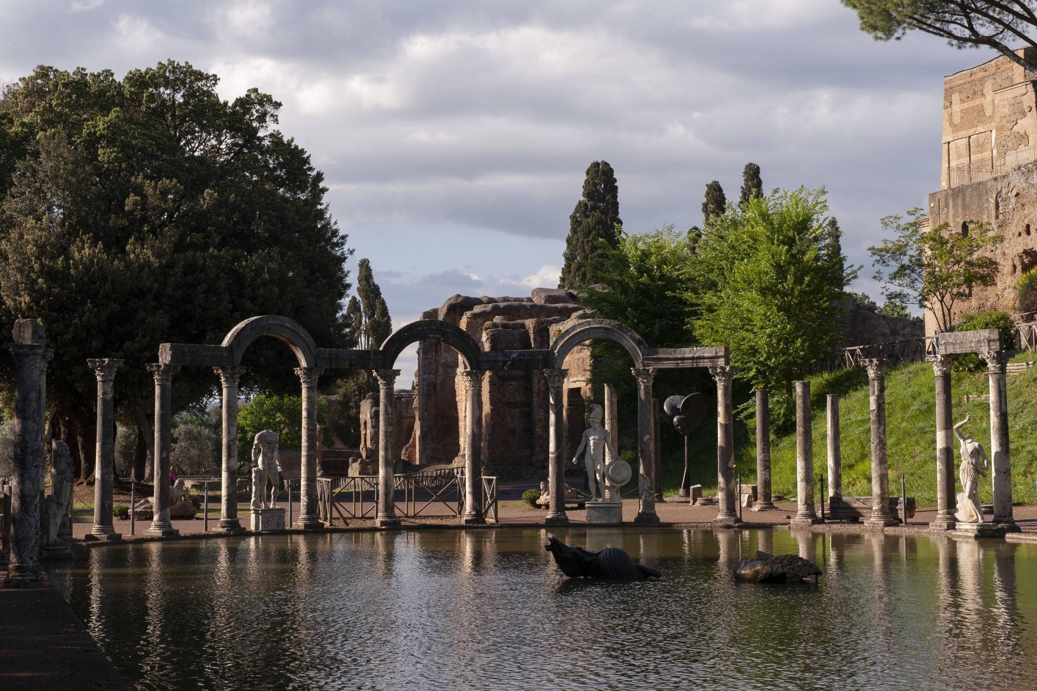 Ancient stone columns and arches stand beside a reflective pond with statues and greenery, under a partly cloudy sky at an archaeological site.