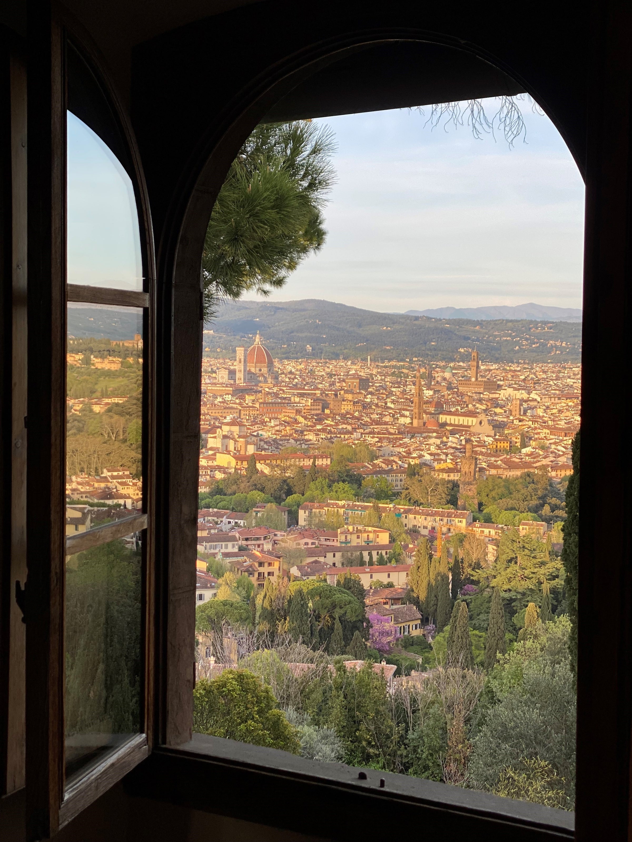 A scenic view of Florence, Italy, framed by an open arched window, showing the city’s historic buildings, the Florence Cathedral’s dome, and lush green trees in the foreground during daylight.