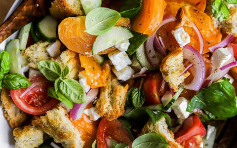 A close-up of a colorful salad with sliced tomatoes, yellow tomatoes, cucumbers, red onions, cubes of feta cheese, fresh basil leaves, and chunks of crusty bread.