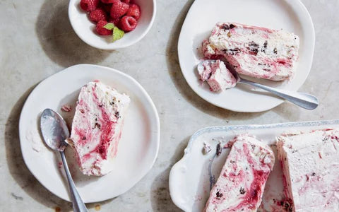 Three plates with slices of raspberry swirl ice cream dessert, each with a spoon. A white bowl of fresh raspberries with a mint leaf sits nearby on a light-colored surface.