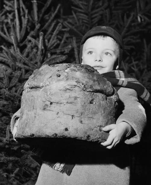 A young child wearing a cap and scarf holds a large round loaf of bread, standing in front of pine trees.