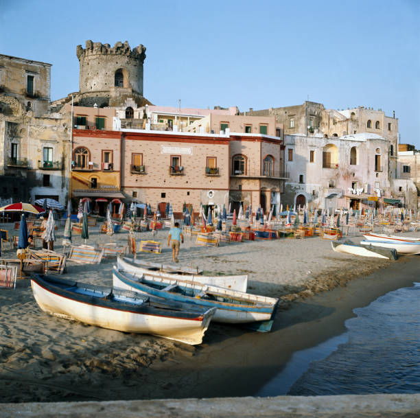 Colorful wooden boats rest on a sandy beach lined with closed umbrellas and lounge chairs, with historic buildings and a stone tower in the background under a clear blue sky.