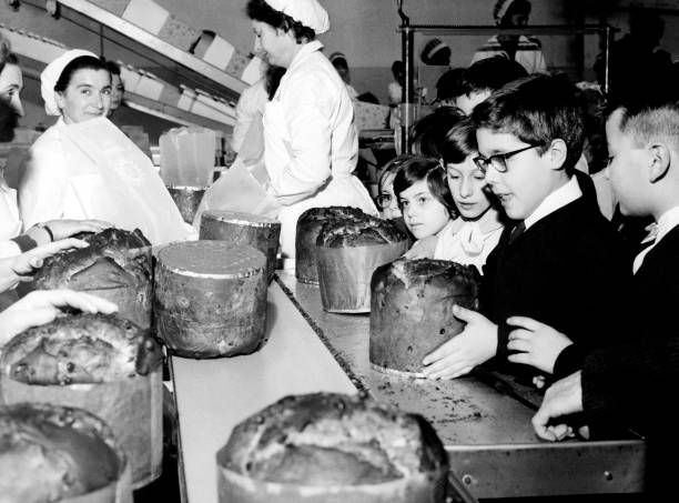Children in a bakery observe workers in uniforms preparing large loaves of bread or cakes on a conveyor belt. The children appear curious and excited as they watch the process.