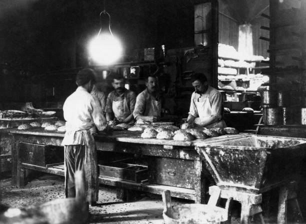 Four bakers work together preparing and shaping loaves of bread on a large wooden table inside a busy, dimly lit bakery, with shelves of bread visible in the background.