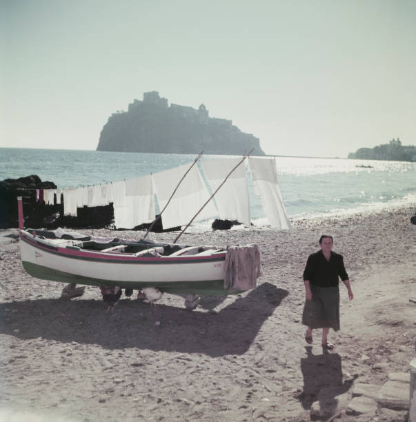 A woman walks on a sandy beach near a boat and laundry hanging to dry. The sea and a large rocky island with a castle are visible in the background under a clear sky.