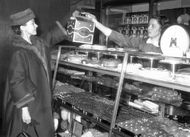 A woman in a fur coat receives a box of pastries from a bakery worker behind a glass display filled with assorted baked goods and chocolates.