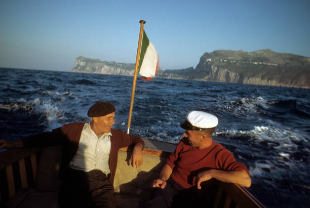 Two men sit and chat on a small boat with an Italian flag, surrounded by choppy blue water and distant rocky cliffs under a clear sky. One man wears a beret, the other a white captains hat.