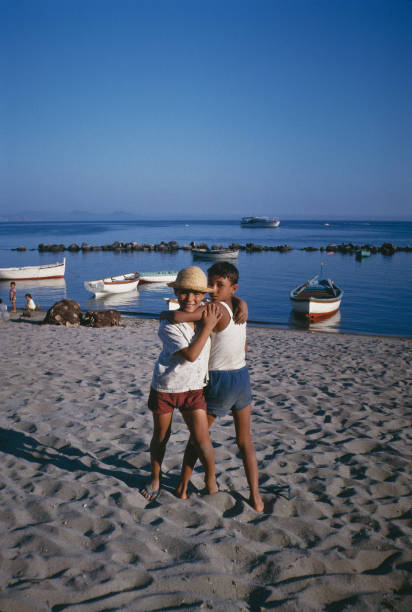 Two children stand hugging and smiling on a sandy beach with boats on calm water in the background under a clear blue sky. Other people and rocks are visible near the shoreline.