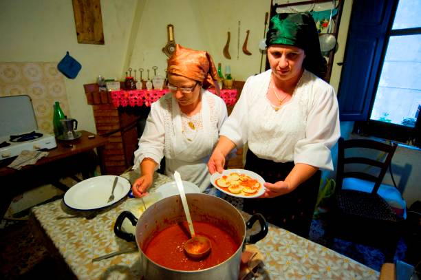 Two women wearing traditional clothing prepare food in a rustic kitchen. One holds a plate of sliced bread with sauce, while the other serves from a large pot of red soup on the table. The room has a cozy, vintage feel.