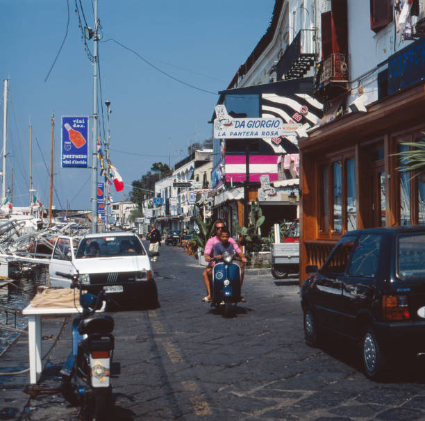 A narrow street by a marina in Italy, with parked cars, scooters, and a couple riding a blue scooter. Colorful buildings, shops, and Italian flags are visible along the bustling street.