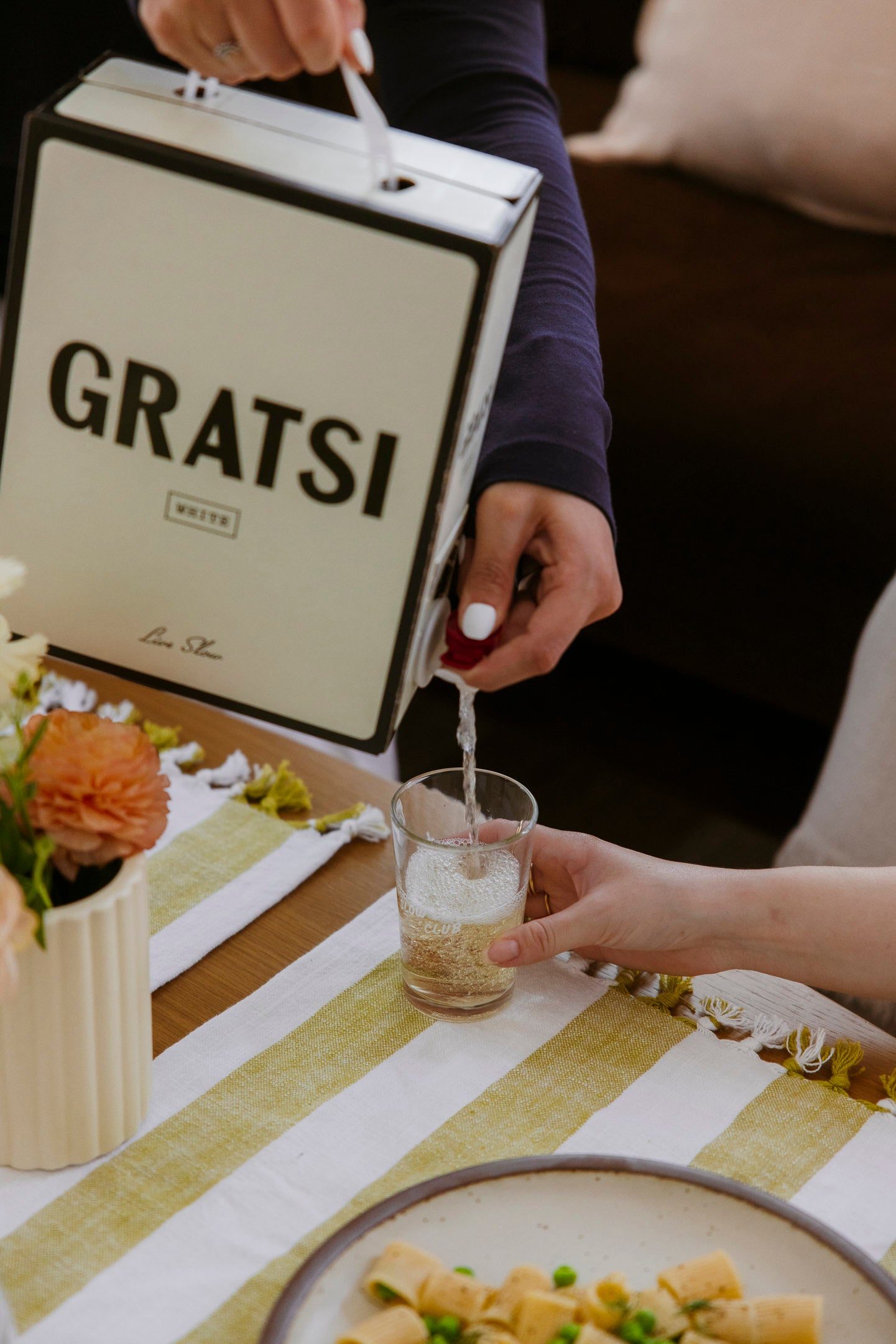 A person pours white wine from a Gratsi box into a glass held by another person at a table set with flowers, a striped table runner, and a plate of pasta.