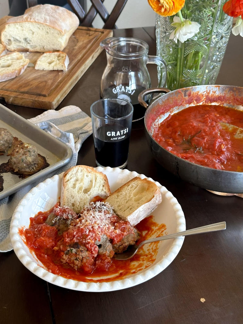 A table set with a bowl of meatballs in tomato sauce, topped with grated cheese and served with bread. Nearby are a tray of bread, a pan of sauce, a glass and pitcher labeled “GRATSI,” and a vase of orange flowers.