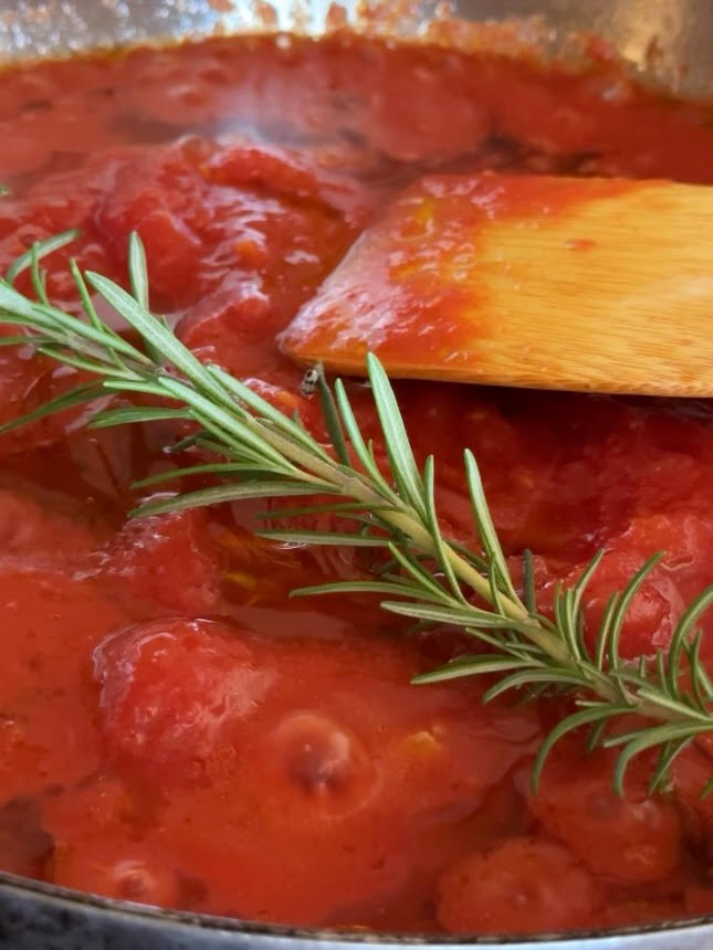 A close-up of tomato sauce simmering in a pan with a wooden spatula and a fresh sprig of rosemary resting on top.