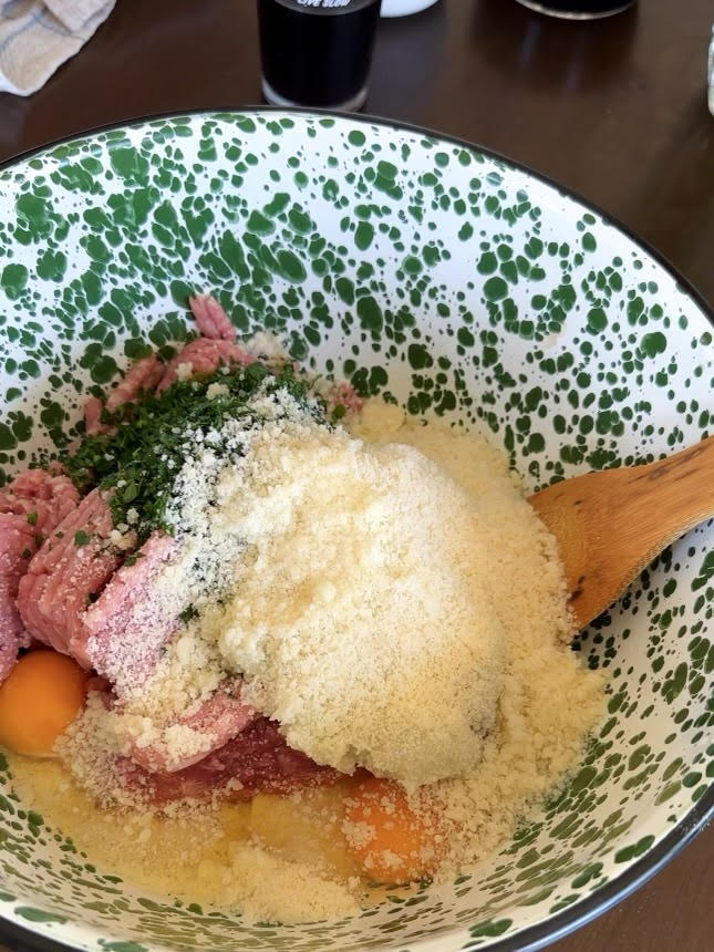 A speckled green and white bowl filled with ground meat, grated cheese, chopped herbs, and raw eggs, being mixed with a wooden spoon.