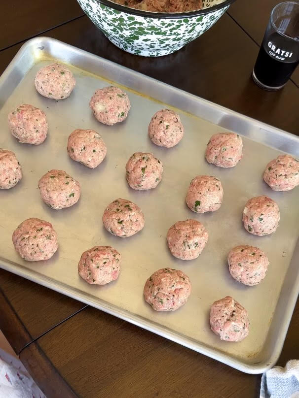 A metal baking sheet with rows of raw, seasoned meatballs on it sits on a wooden table. In the background, there is a bowl with more meat mixture and a glass of dark liquid.