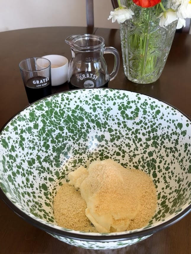 A green and white speckled bowl with butter and breadcrumbs inside sits on a wooden table. In the background are a glass pitcher, a glass, a small container, and a vase of flowers.