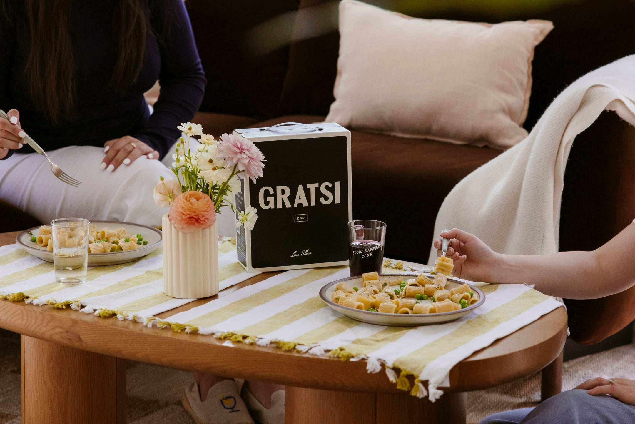 Two people sit at a wooden table with plates of pasta, a GRATS! wine box, glasses of drinks, and a vase of flowers on a striped table runner. The scene is cozy, casual, and indoors.
