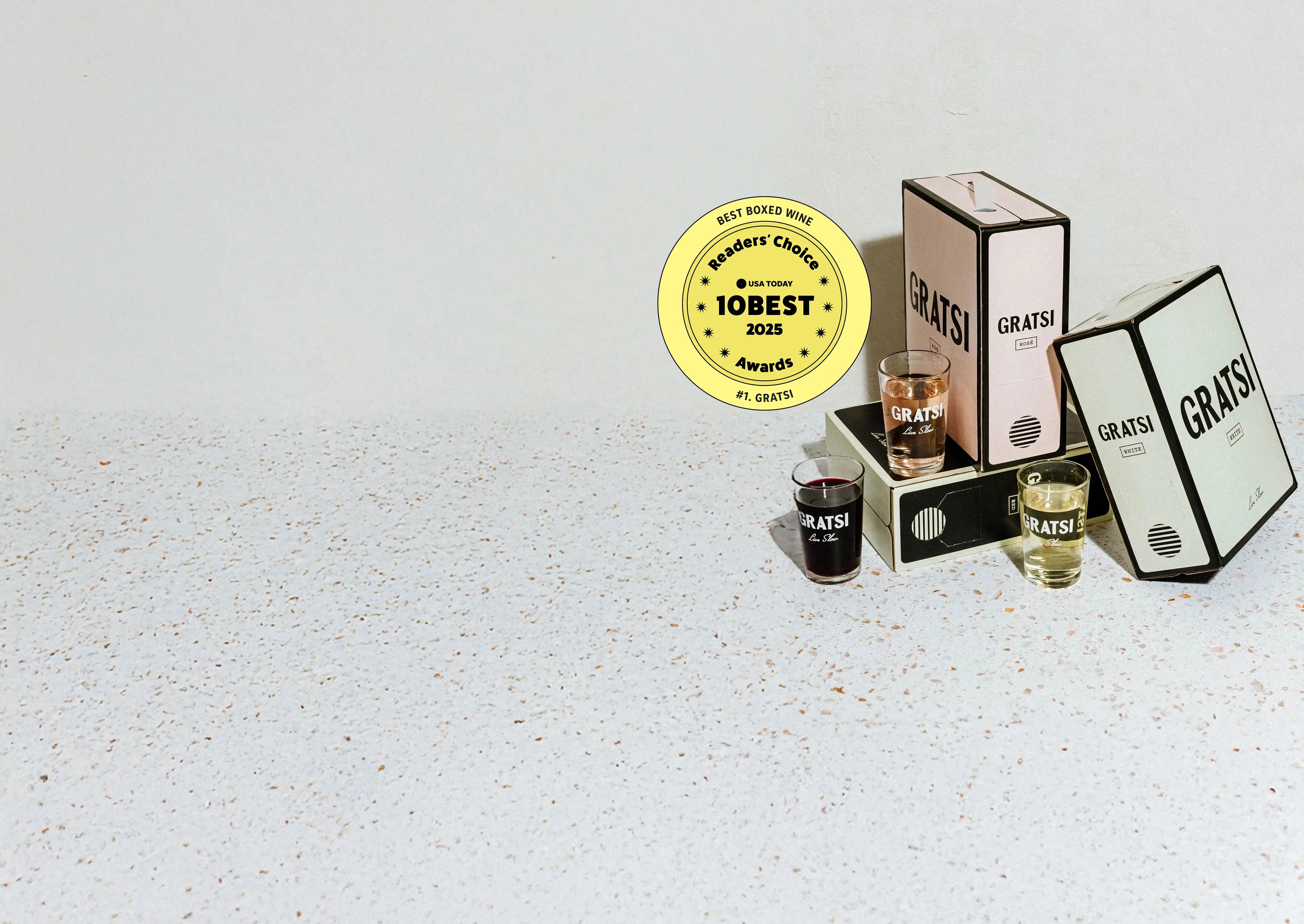 Three Gratsi boxed wine packages in pink, white, and gray with matching glasses are displayed on a speckled surface. A gold 10 Best 2023 badge for Best Boxed Wine is shown above the boxes.
