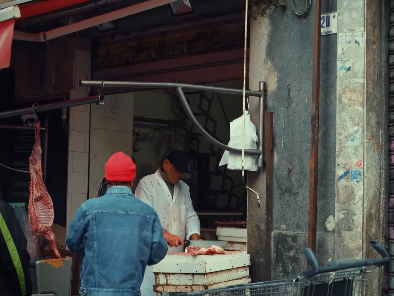 A butcher in a white coat and dark cap is cutting meat at an outdoor market stall. A person in a red beanie and denim jacket stands nearby. The scene is set on a weathered city street.