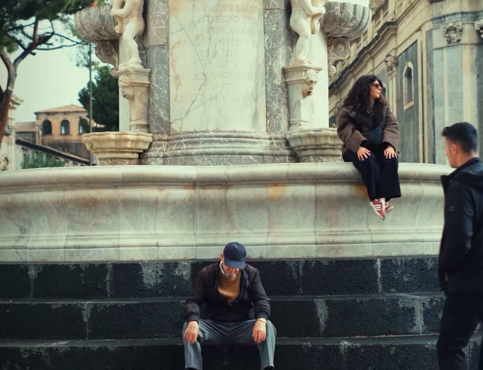 A man sits on the ground in front of a large stone fountain, while a woman wearing sunglasses sits on the fountain ledge above him. Another person walks past them.