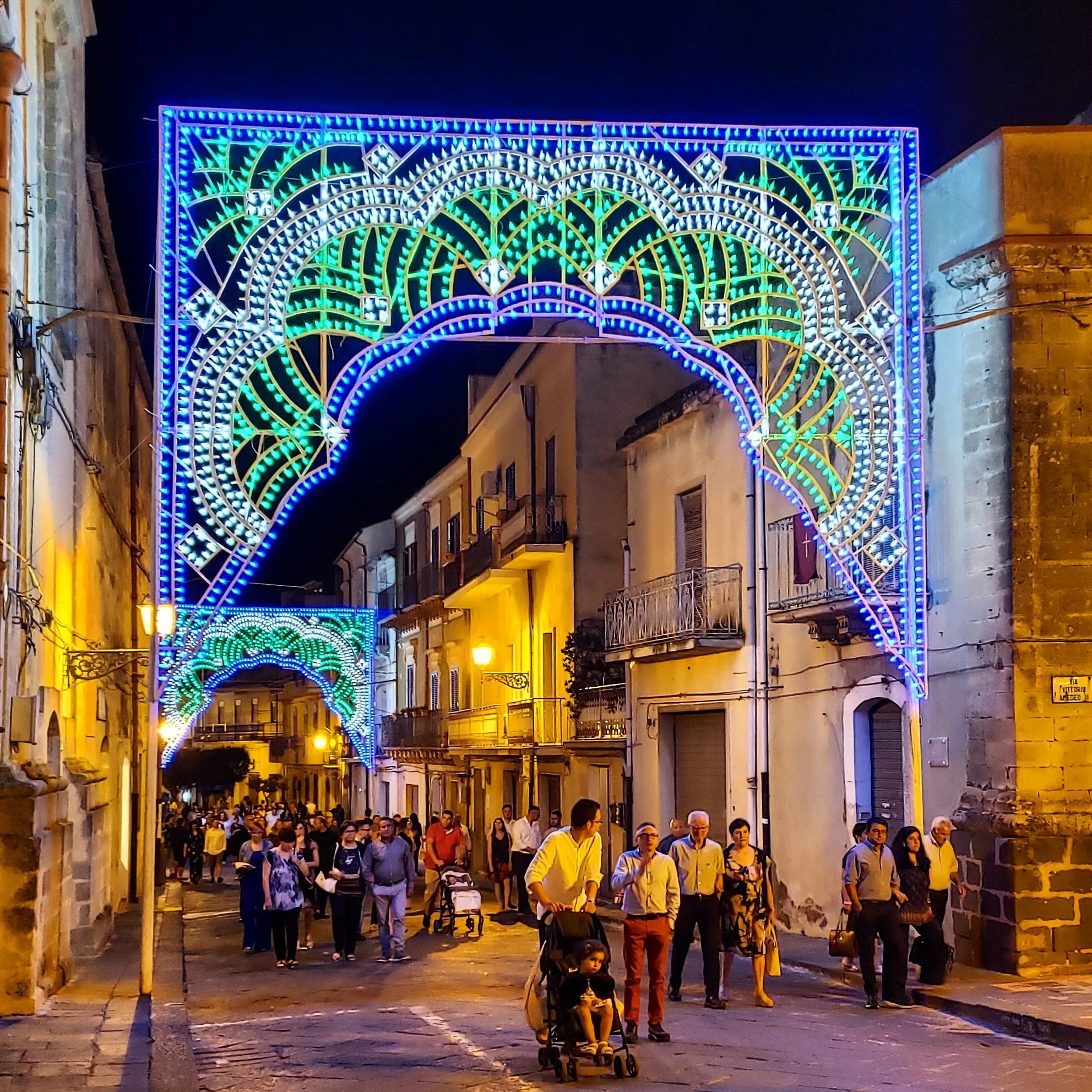 A lively street scene at night shows people walking under brightly lit, colorful archways decorated with green and blue lights in an old town with stone buildings. The festive atmosphere suggests a local celebration or festival.