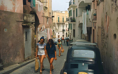Two women walk up a narrow, sunlit street lined with parked cars and old buildings, while another woman and a child walk behind them. Laundry hangs on lines and balconies above the street.