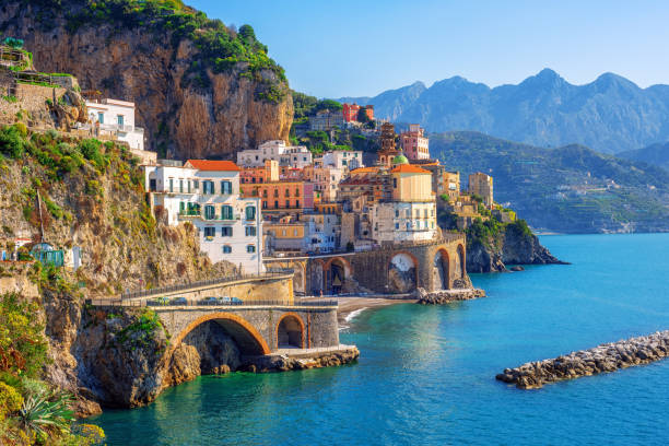 Colorful buildings sit atop dramatic cliffs beside the blue sea on the Amalfi Coast in Italy, with arched bridges and rocky mountains in the background under a clear sky.