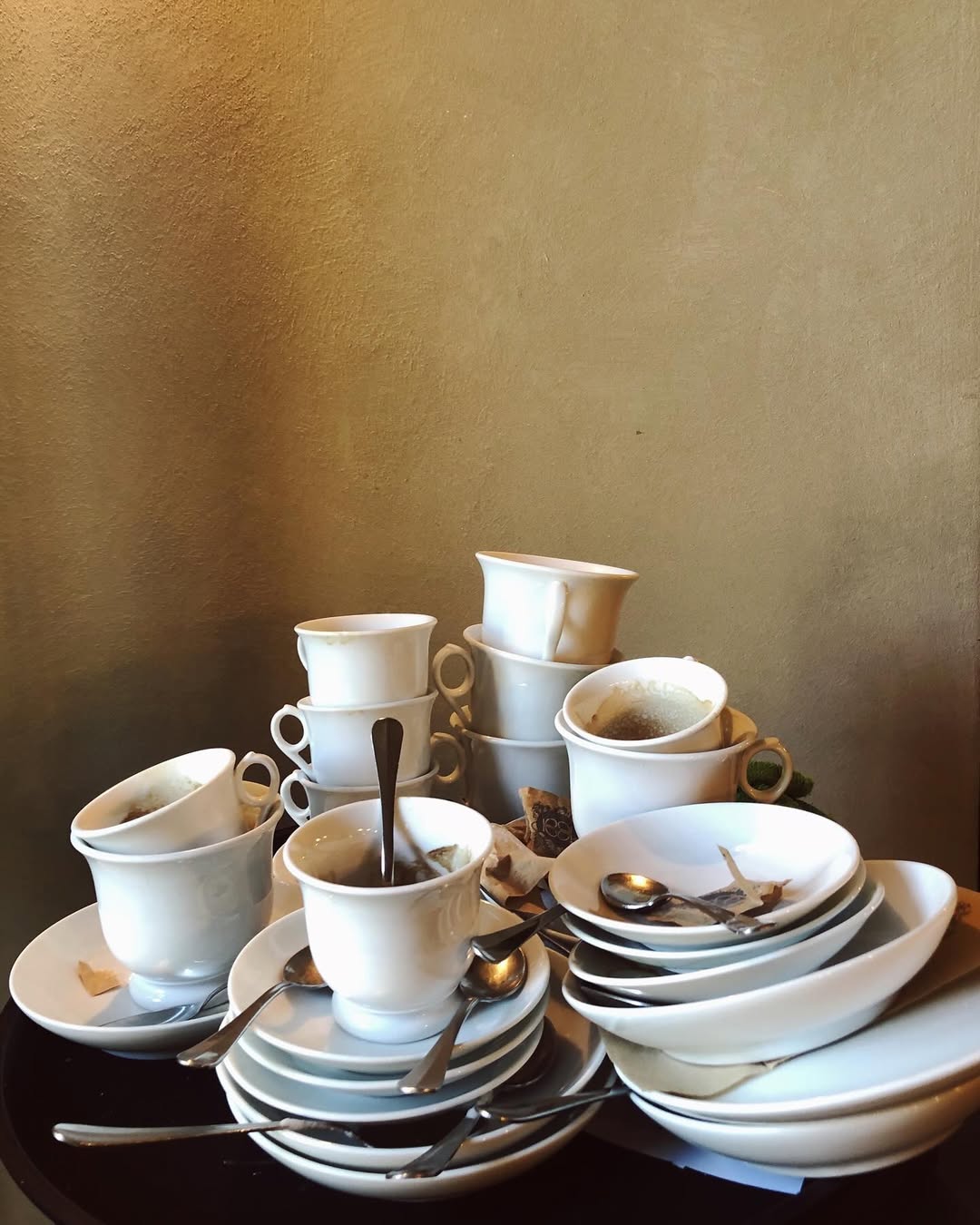 A stack of used white cups, saucers, and plates with several teaspoons and used tea bags on a dark table against a plain beige wall.