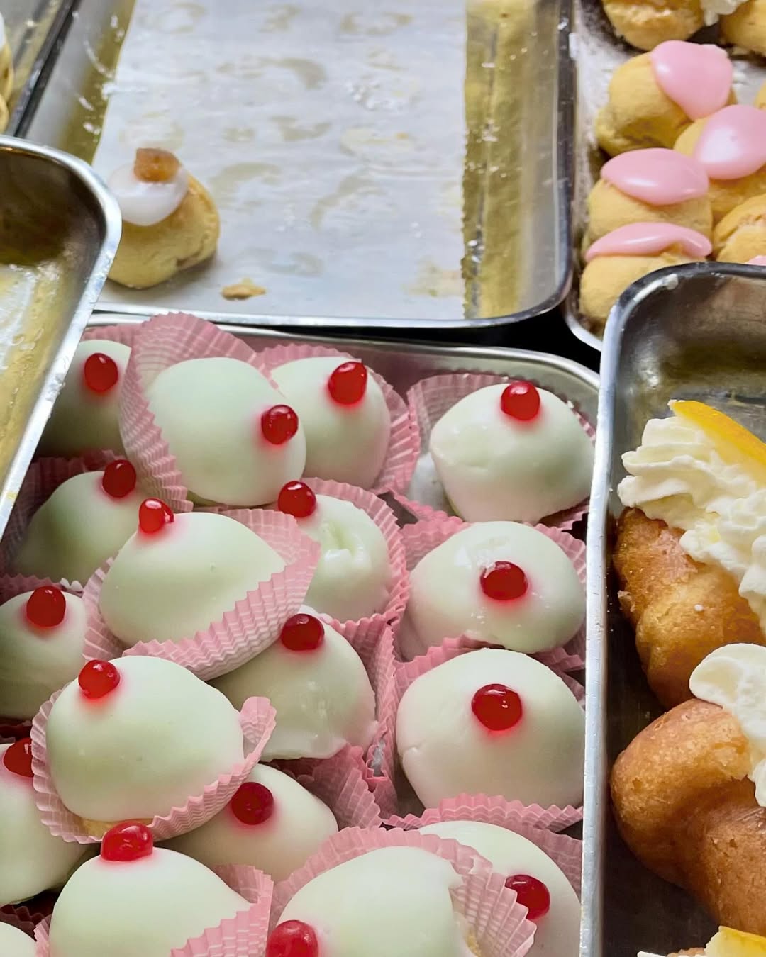 Assorted pastries on metal trays, including white dome-shaped treats with red cherries on top in pink wrappers, and other pastries decorated with cream and pink or orange toppings.