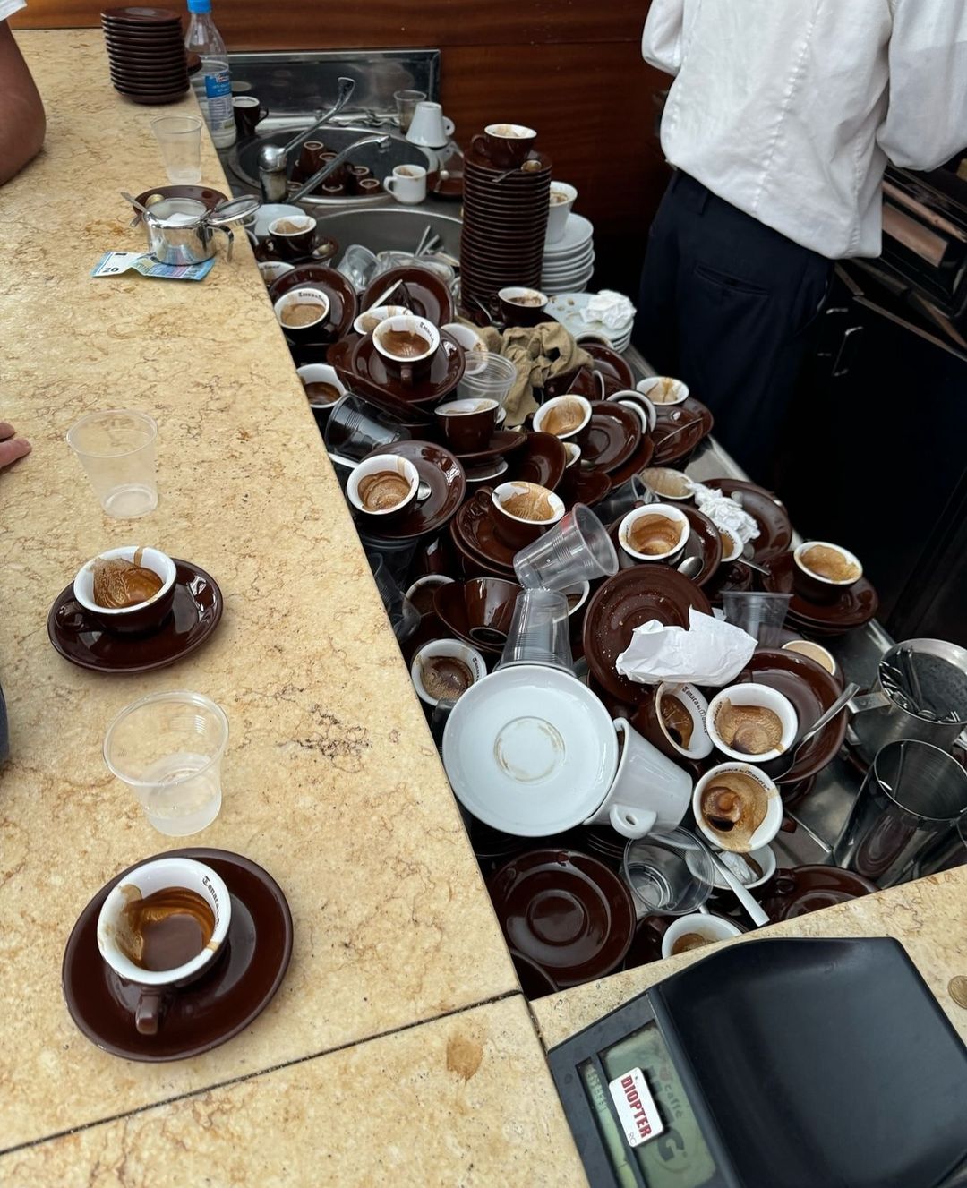 A large pile of empty espresso cups and saucers is stacked messily behind a counter, with used cups and plastic glasses scattered on the countertop. A person in a white shirt stands nearby.