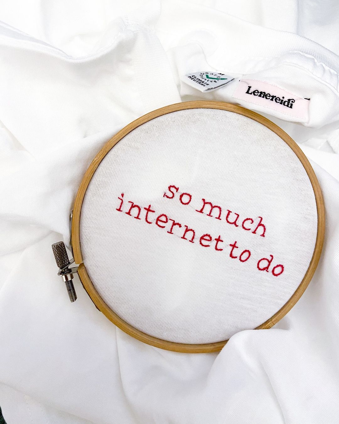 An embroidery hoop with white fabric features red stitched text reading so much internet to do, resting on a white cloth. A clothing tag reading Leneredi is partially visible in the background.