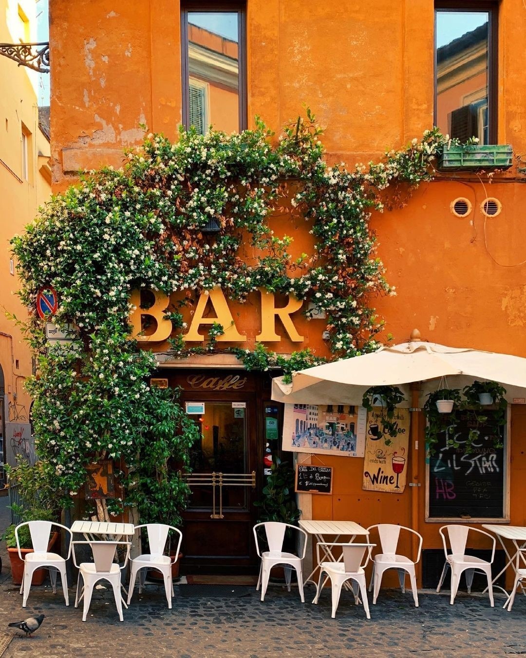 A charming orange building with a flower-covered entrance labeled BAR, white chairs and tables outside, and a white umbrella. The scene is set in a cobblestone street with warm, inviting light.
