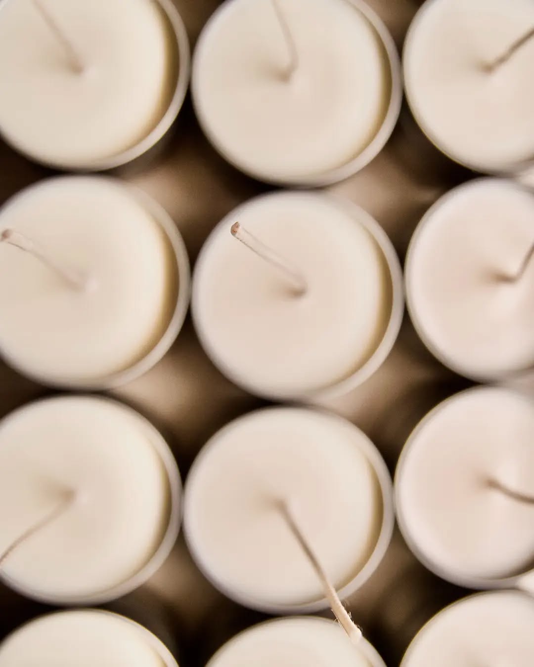 Close-up, top-down view of several white candles arranged closely together in a neat grid, with their wicks centered and pointing upwards.