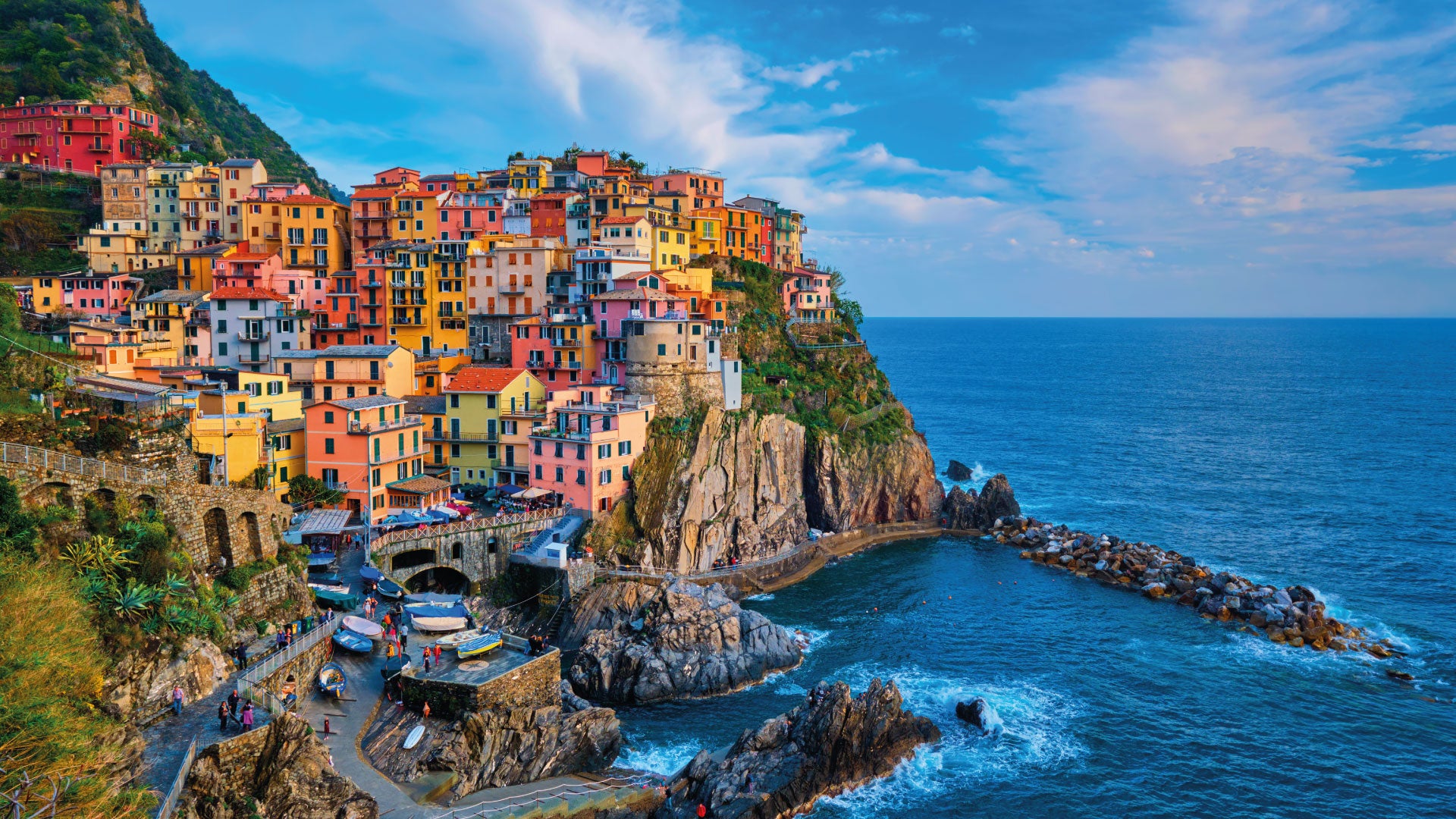 Colorful buildings of the village of Manarola in Cinque Terre, Italy, sit atop a rocky cliff overlooking the blue waters of the Ligurian Sea under a partly cloudy sky.