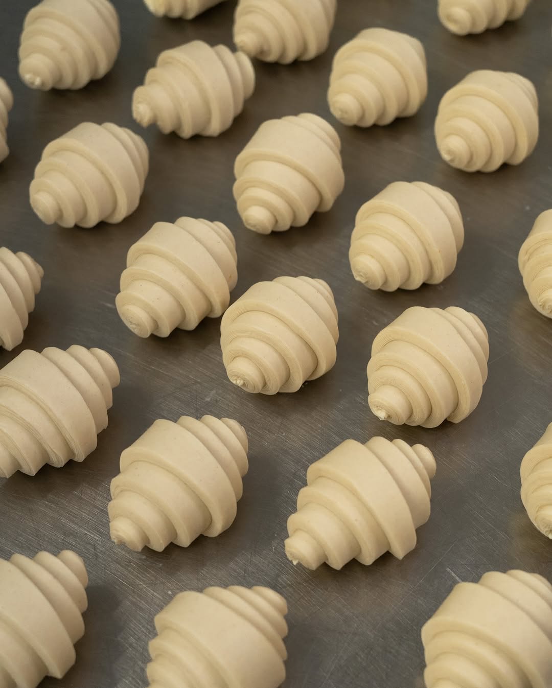 Rows of unbaked, neatly rolled croissant dough spirals are arranged evenly on a metal baking sheet, ready for baking.