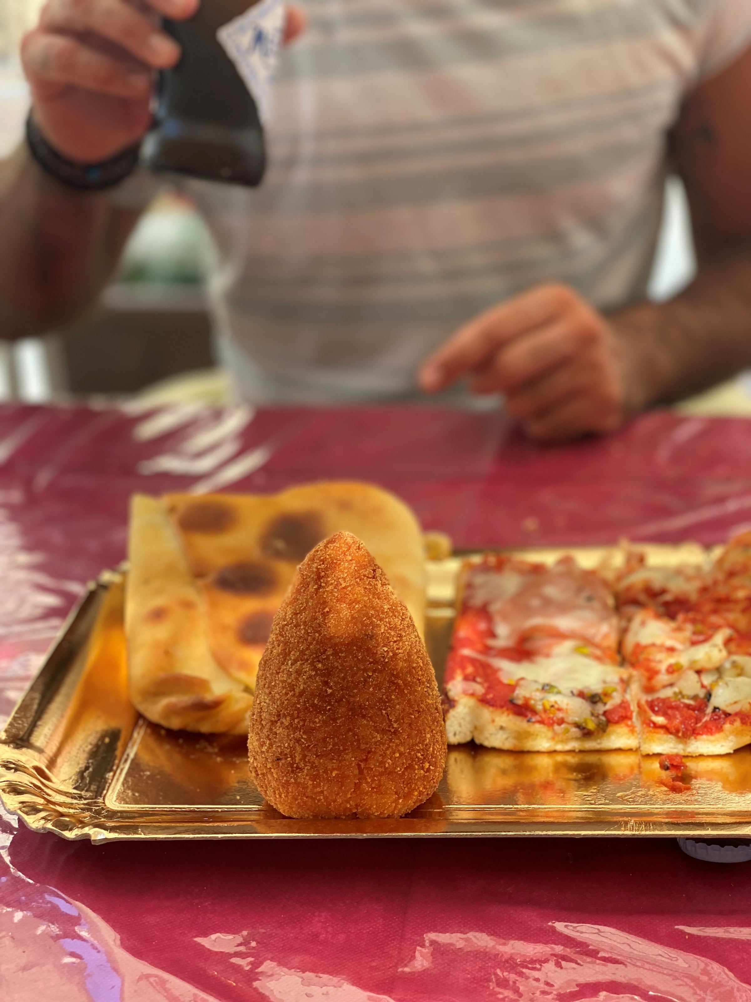 A person sits at a table with a tray of Italian food, including a golden arancino, slices of pizza, and focaccia. The person holds a phone, and the table is covered with a red tablecloth.