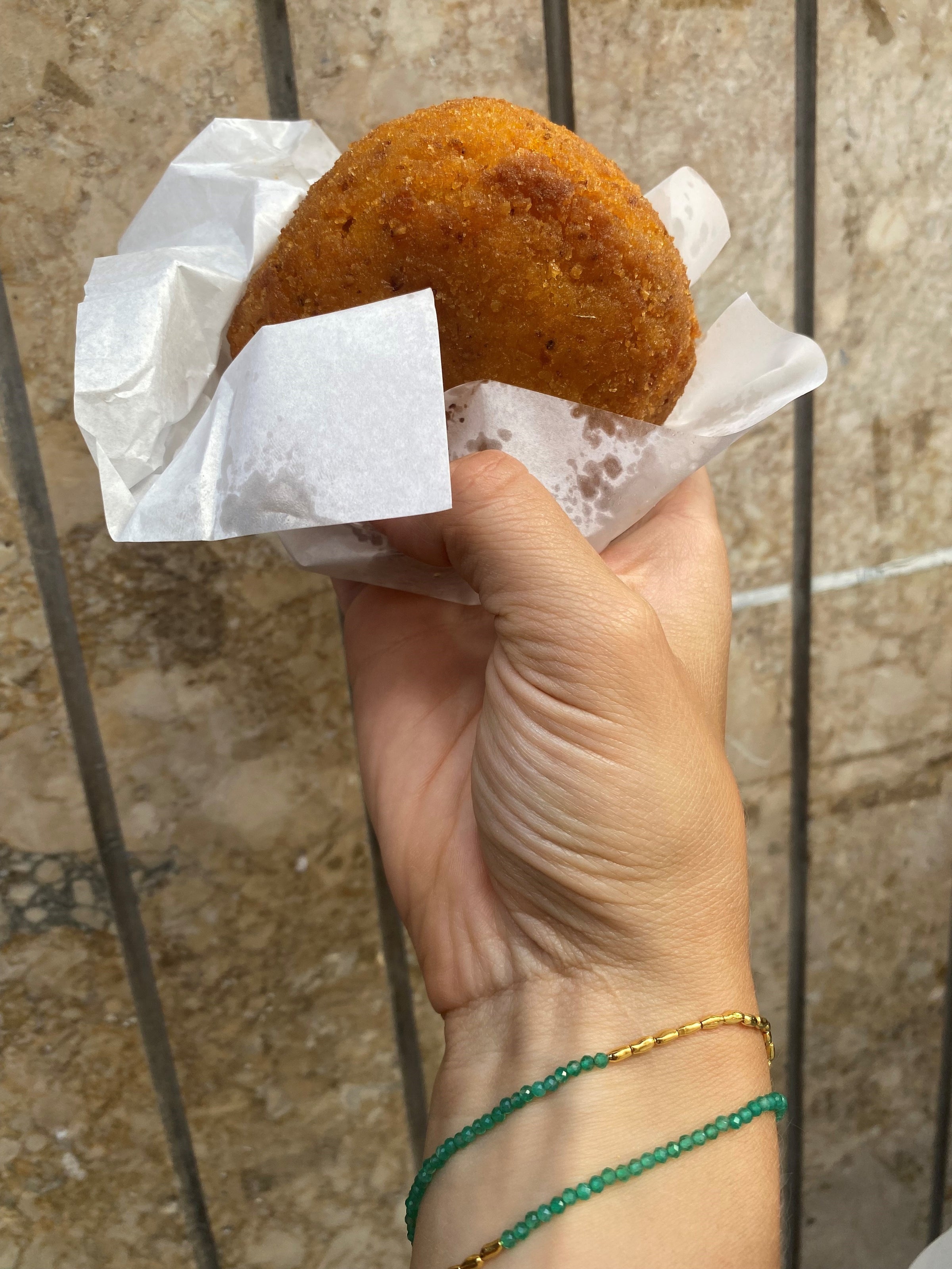 A hand with green and gold bracelets holds a round, golden-brown fried snack in white paper against a tiled wall background.