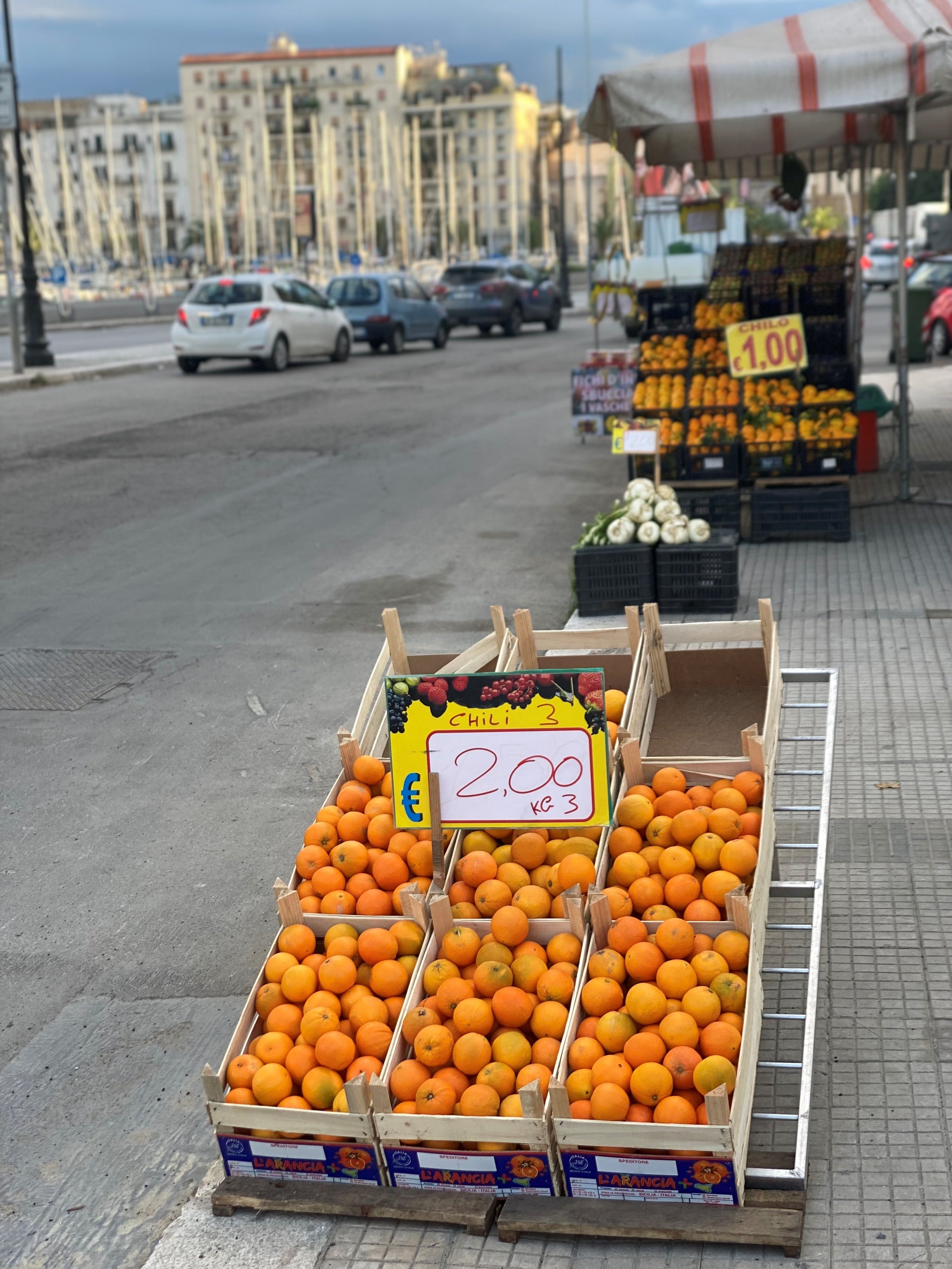Crates of oranges labeled 2.00 €/kg displayed at a sidewalk fruit stand near a street, with more produce and buildings visible in the background.