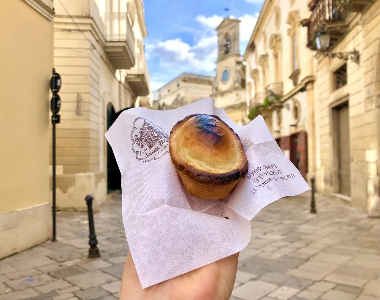 A hand holds a small pastry wrapped in white paper on a cobblestone street lined with old buildings under a blue sky with scattered clouds. A clock tower is visible in the background.