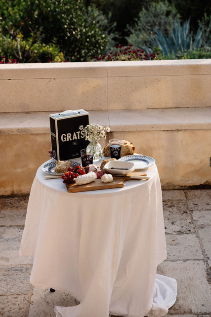 A round table with a white cloth displays a cheese board with assorted cheeses, grapes, bread, two glasses of Red Wine by Gratsi, a Gratsi wine box, and a vase of small white flowers on an outdoor stone patio.