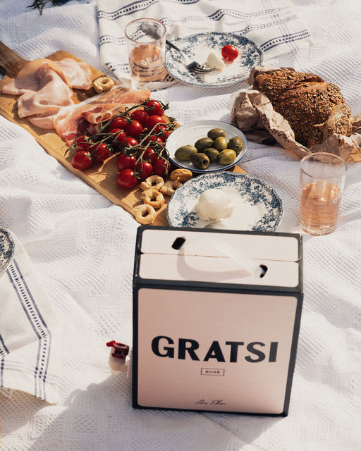 A picnic spread on a white blanket features bread, olives, cherry tomatoes, cold cuts, and glasses of Gratsi Rosé Wine (zero sugar), all surrounded by blue and white plates and linens.